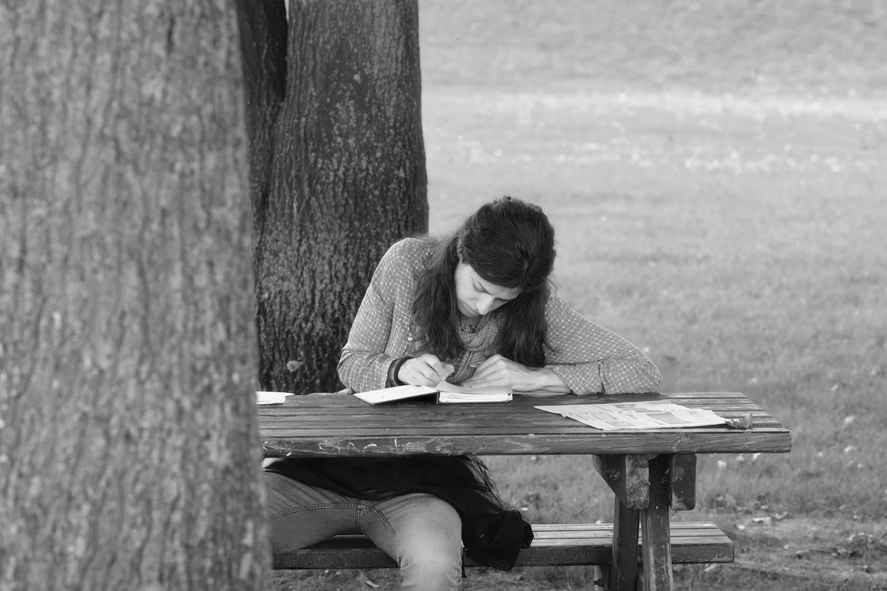 ragazza giovane intenta a scrivere a mano in un parco seduta sotto un albero e su un tavolino da pic nic