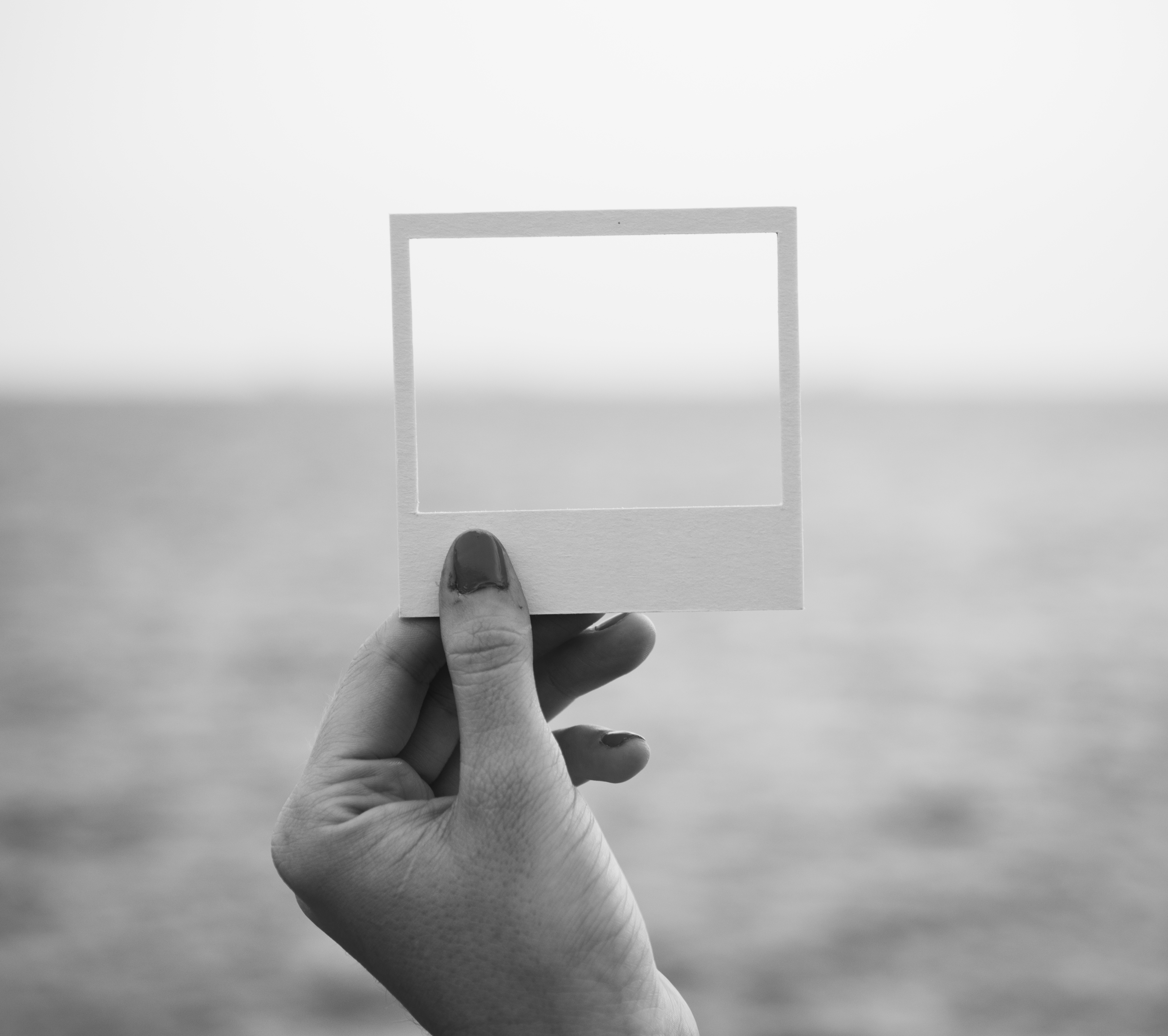 una mano femminile tiene tra le dita la cornice di una polaroid senza foto e sullo sfondo il mare